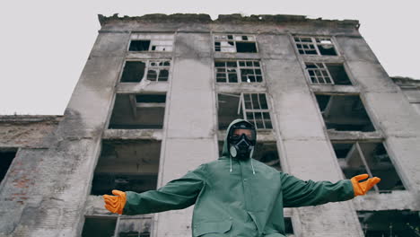 abandoned place and a person in safety suit. survivor in gas mask and hazmat suit standing with stretched arms on ruined building background. chemical catastrophe.