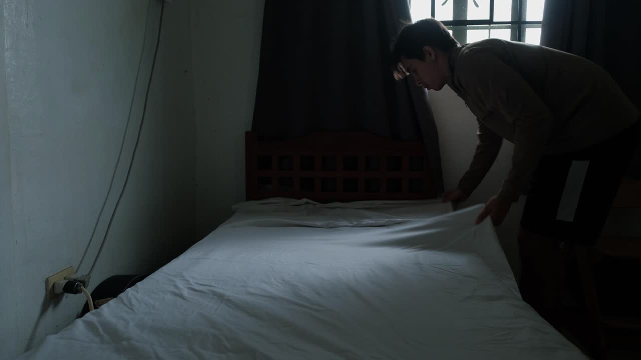 Young man fixing bed sheets on twin size bed in sunlit bedroom -static wide shot