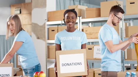 voluntaria afroamericana sosteniendo cajas de donación y mirando la cámara en un almacén de caridad mientras sus compañeros de trabajo trabajan en cajas de embalaje