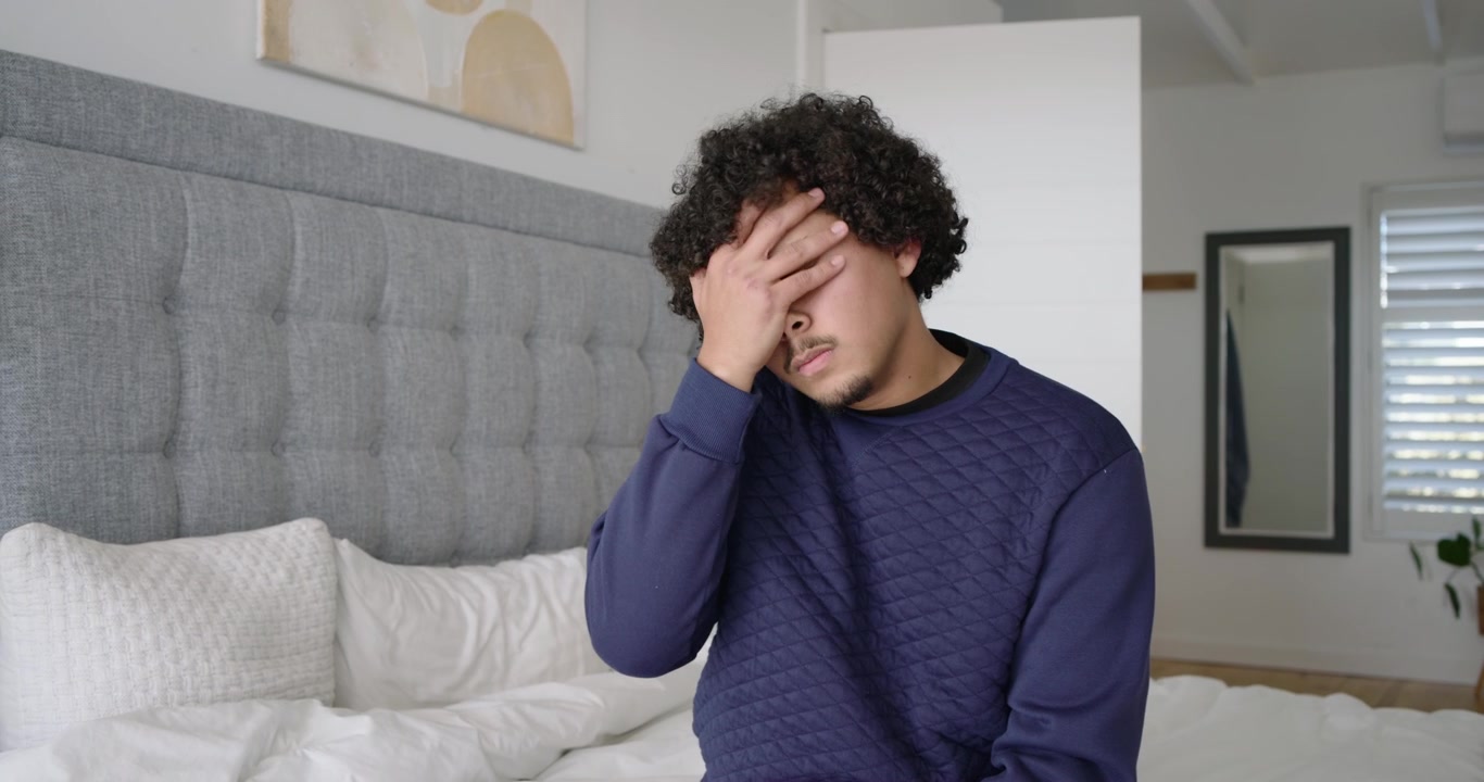 Coppia afroamericana che litiga, la donna gesticola e se ne va, l'uomo seduto sul letto sospira coprendosi gli occhi.