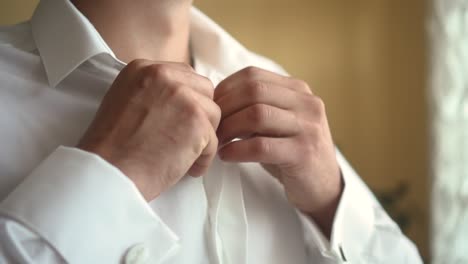 man buttons up his white shirt standing in the front of a bright window 01