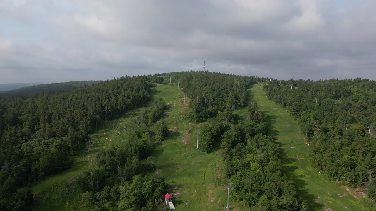 Aerial footage flying up thick green tree covered mountain following chairlift on cloudy, overcast summer day. Calabogue Ontario
