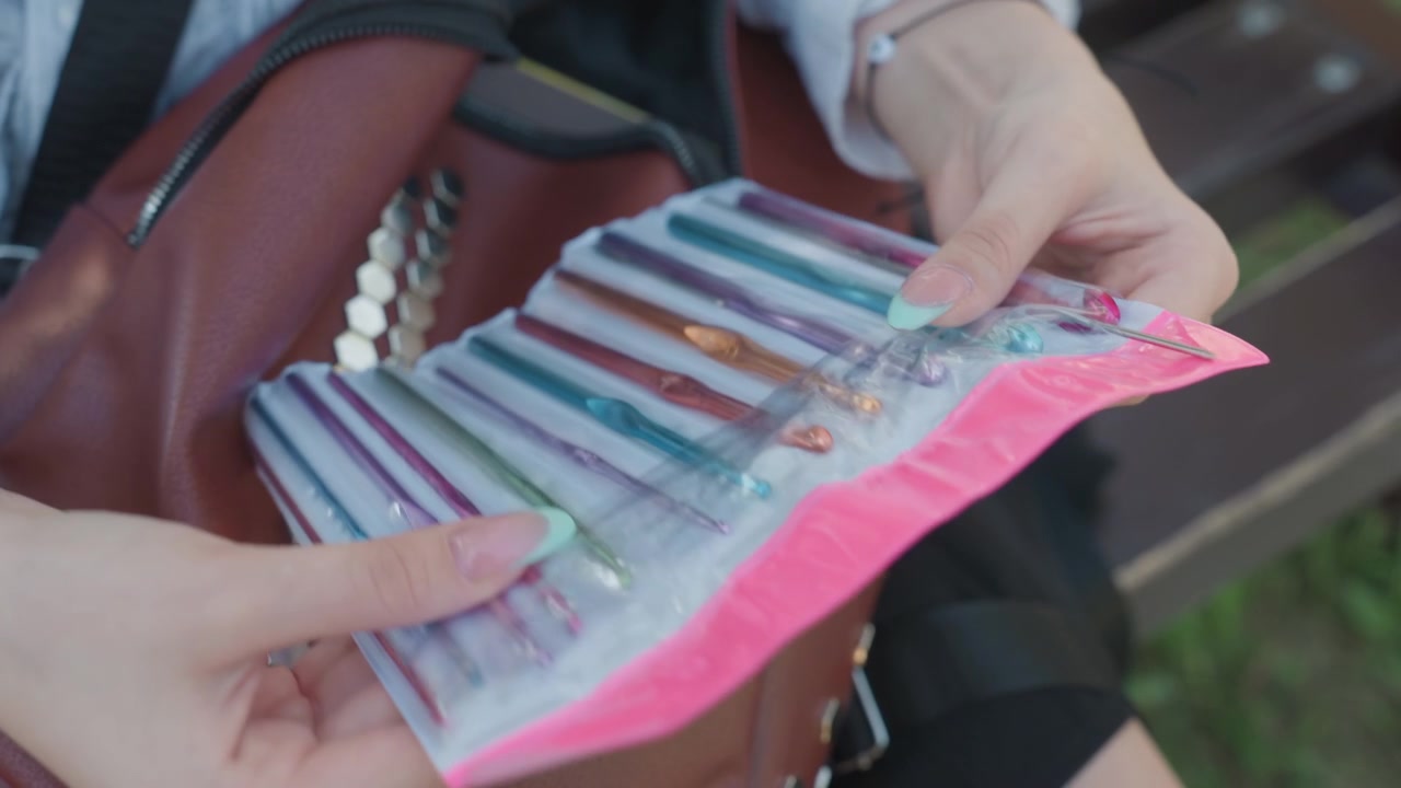 Primo piano di una giovane donna con smalto azzurro che maneggia uncinetti colorati da una confezione rosa mentre è seduta su una panchina all'aperto. La donna esamina gli strumenti rivelando varie dimensioni e colori