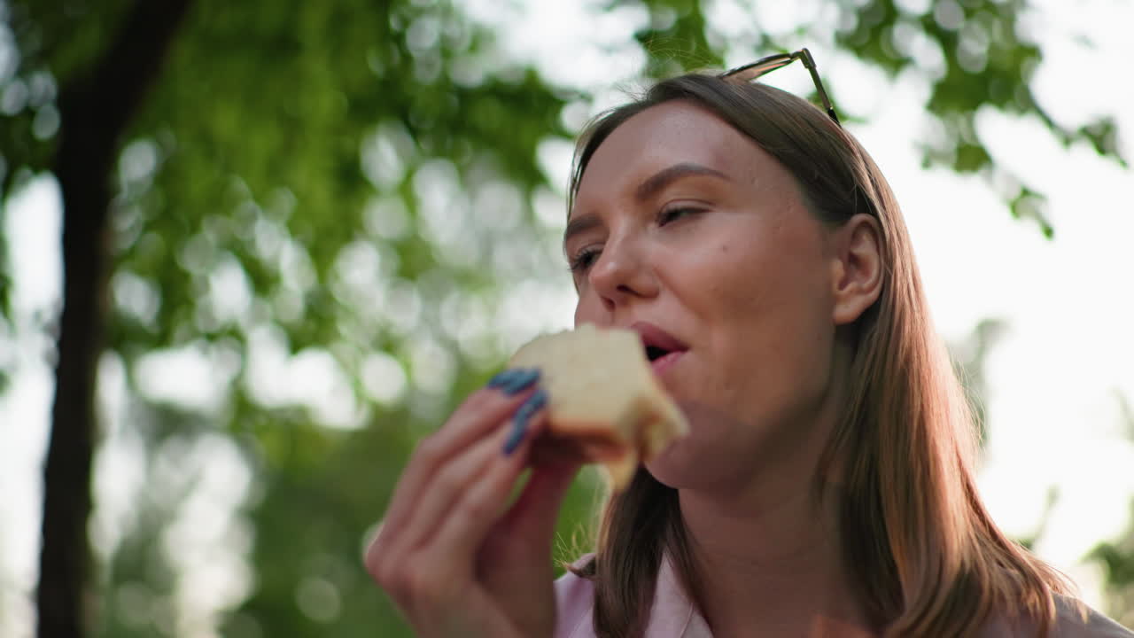 mujer con una camiseta rosa con gafas de sol descansando en la cabeza disfrutando de un sándwich a la parrilla con vegetación borrosa en el fondo, al aire libre con luz natural