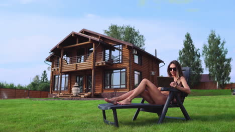 una hermosa mujer delgada yace en una silla larga en un traje de baño con una tableta en el fondo de una gran casa de campo con gafas de sol y baños de sol en el sol viendo fotos en las redes sociales