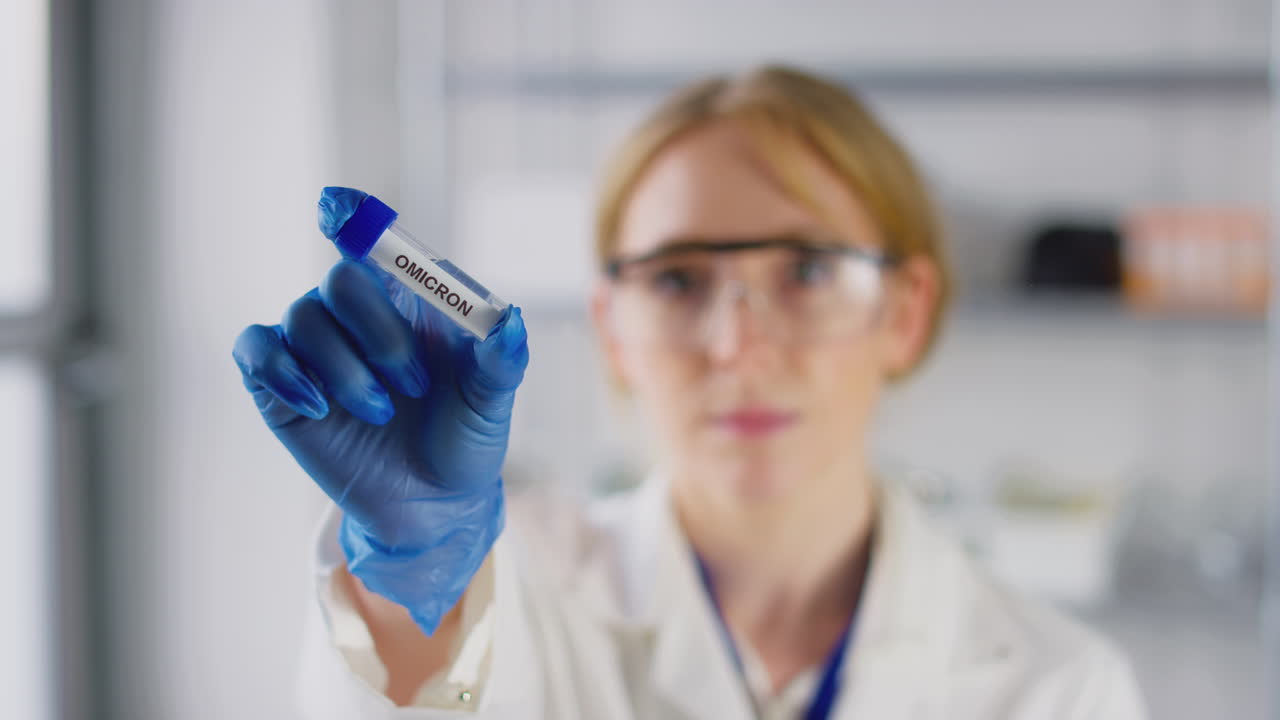 Female Lab Research Worker Wearing Safety Glasses Holding Test Tube Labelled Omicron
