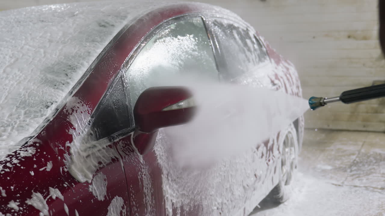Side view of red car covered in soap foam being rinsed with high pressure water spray by someone in garage interior showing dynamic flow of water and dripping soap suds during professional cleaning