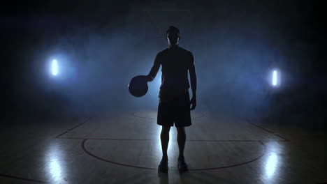 Der-Basketballspieler-Steht-Auf-Einem-Dunklen-Spielplatz-Und-Hält-Den-Ball-In-Seinen-Händen-Und-Blickt-In-Die-Kamera-Im-Dunkeln-Mit-Hintergrundbeleuchtung-In-Zeitlupe-Und-Um-Rauch-Herum