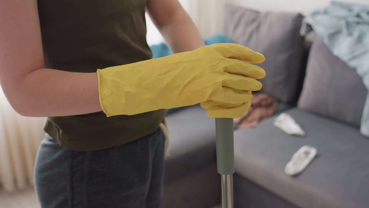 Side view of cleaner wearing yellow gloves holding mop handle with both hands beside couch with scattered clothes showing daily cleaning routine and household