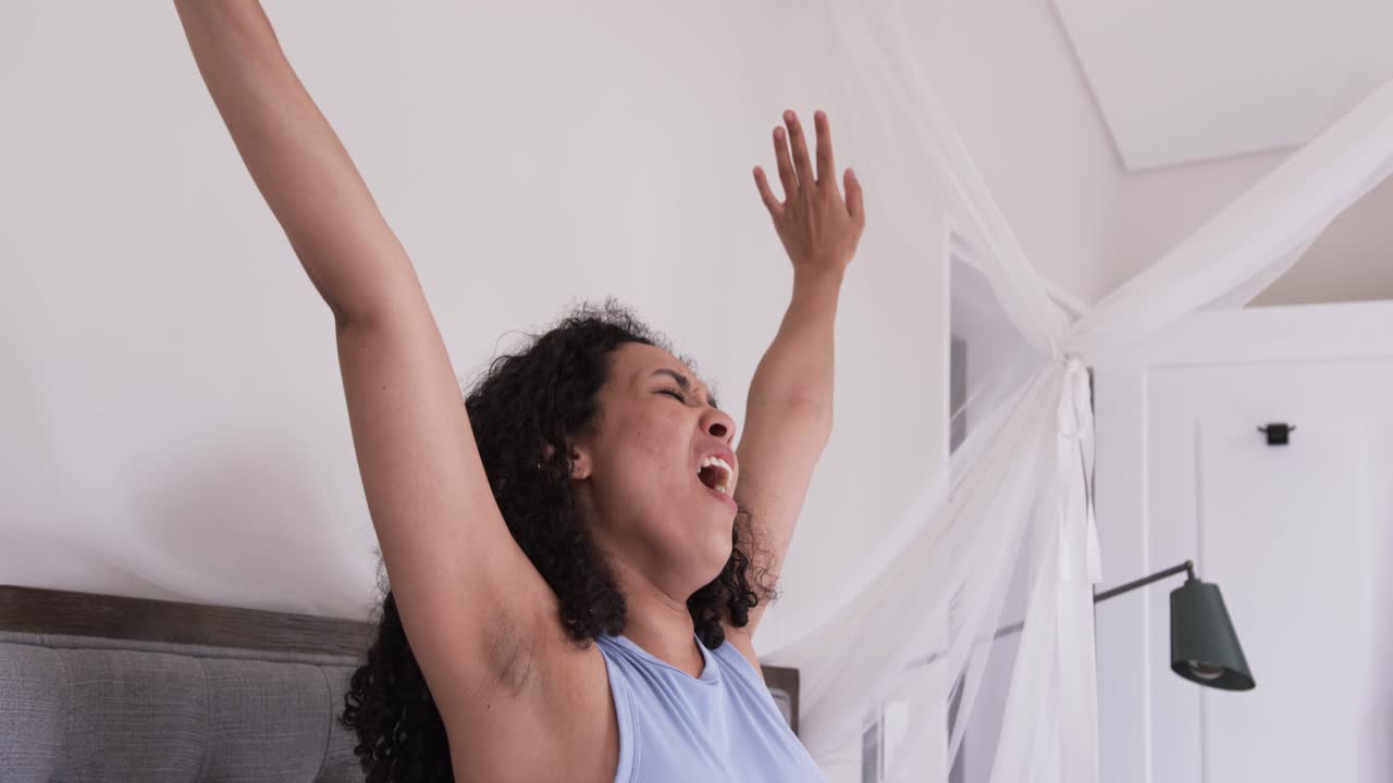 La luce del mattino sveglia una donna, seduta sul letto, che si massaggia il collo e allunga le braccia sopra la testa per svegliarsi.