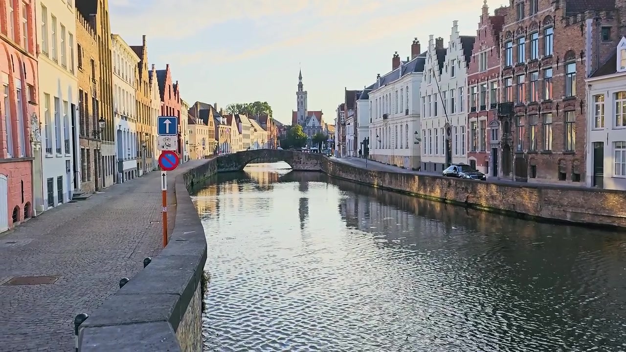 Canale storico fiancheggiato da vecchi edifici in mattoni, strada acciottolata, ponte in pietra e torre di chiesa riflessa nell'acqua calma, una pacifica scena di lungomare europeo nella morbida luce della sera.