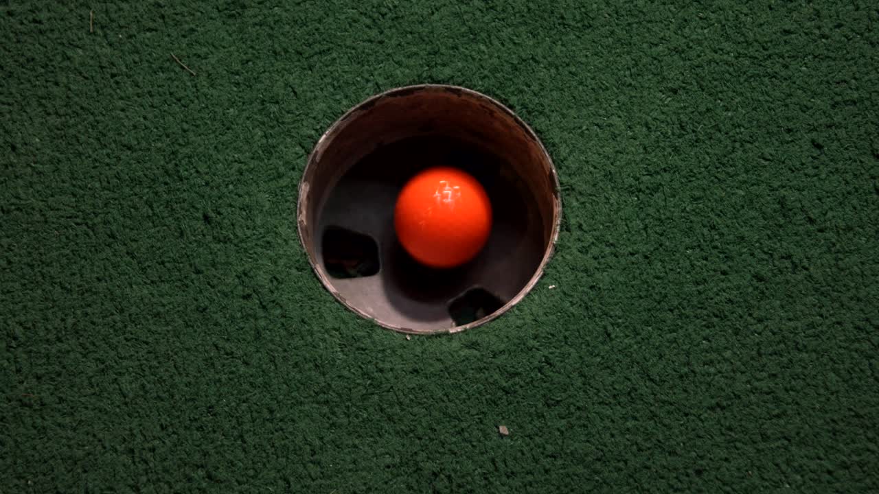 A close up birds eye view of an orange mini golf ball falls into the golf hole and bounces in the pocket on a course