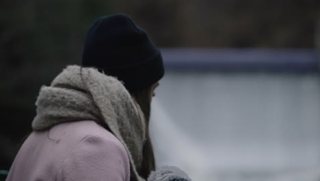 a young woman in winter suit standing in a bridge watching the rushing waters flowing through coaticook river dam in quebec, canada - rack focus, slow motion