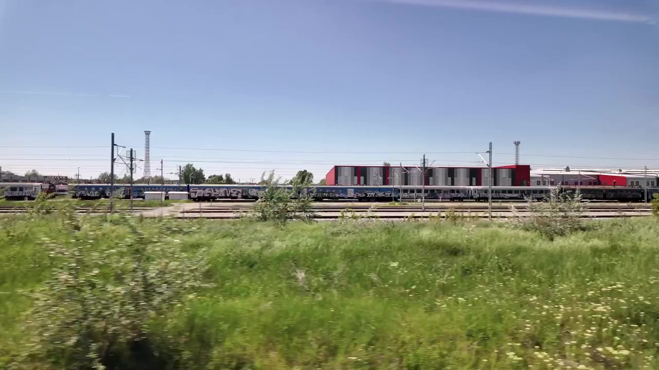Tog er parkeret langs sporene i et travlt jernbaneområde. Græs og små planter vokser nær sporene. Kommunikationstårne og bygninger er synlige i det fjerne.