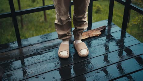 Close-up-shot:-a-person-in-slippers-stands-on-a-floor-of-dark-boards-during-the-rain-outside