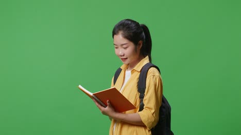 side view of asian woman student with a backpack holding and reading a book while standing in the green screen background studio