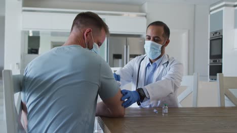 Mixed-race-male-doctor-wearing-face-mask-giving-vaccine-to-male-patient-at-home