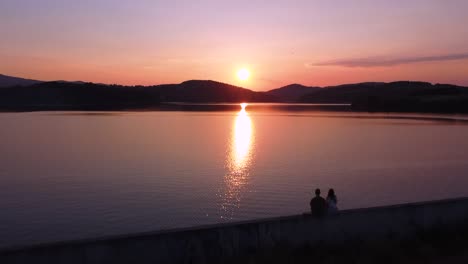 Paar-Sitzt-Bei-Sonnenuntergang-Auf-Der-Mauer-Am-See