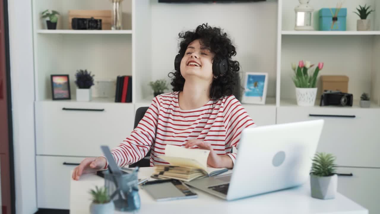 retrato de una chica rizada y bonita sentada en un escritorio en una oficina luminosa, riéndose con una hermosa sonrisa en auriculares inalámbricos