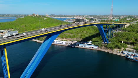 Drone-rises-above-Queen-Juliana-Bridge,-Willemstad-Curacao-as-cars-drive-by-on-sunny-day