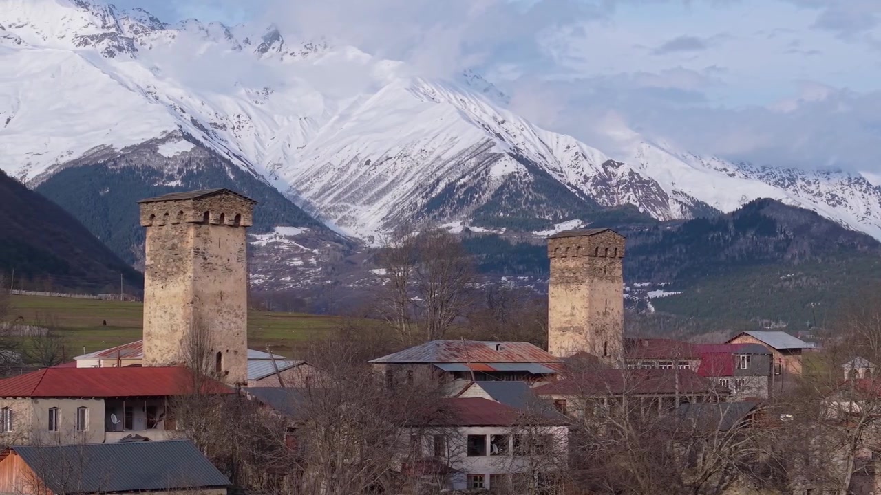 Imagens de drone mostrando casas tradicionais Svan e uma torre Svan medieval, emolduradas por vales verdejantes e os majestosos picos nevados do Cáucaso