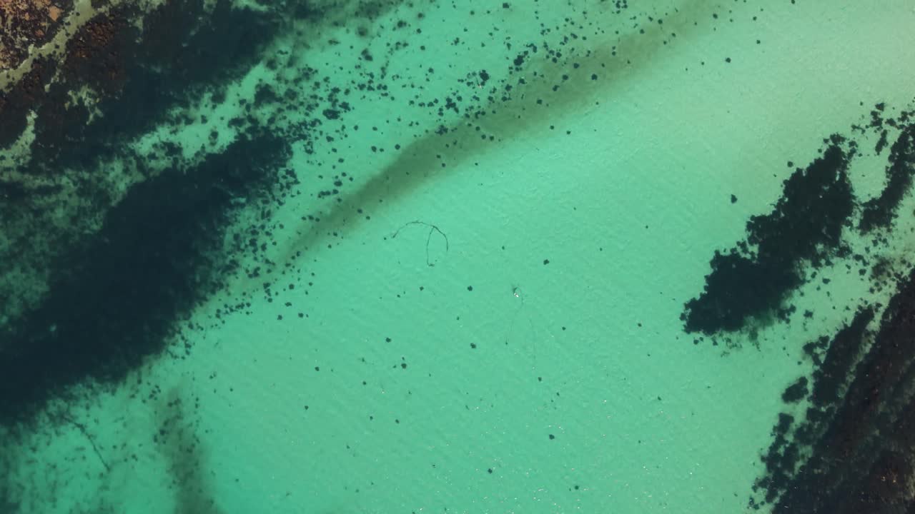 High drone footage over totally clear waters of bay seeing to the seabed unhindered with boats at anchor flying slowly towards rocky shoreline in bright sunshine