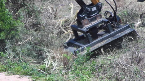 Close-up-shot-on-a-windy-day-in-slow-motion-of-a-yellow-and-black-plant-machinery-arm-with-a-chain,-cutting-through-and-weeding-some-semi-dry-bushes,-leaves-and-branches-in-Spain