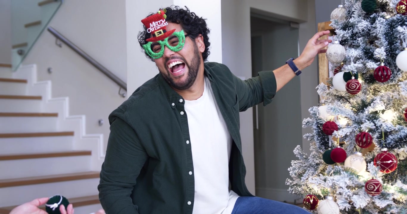 African American man reaching, decorating tree with bauble by stairs donning Merry glasses smiling