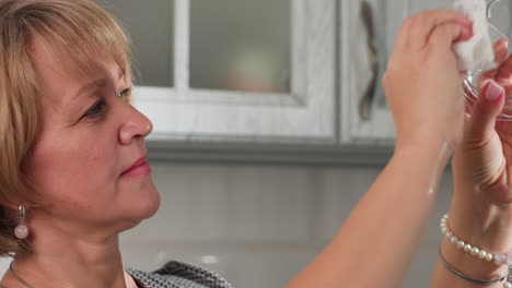 young lady carefully drying transparent glass cup using tissue paper while inspecting for spots and cleanliness in kitchen with cabinets in background