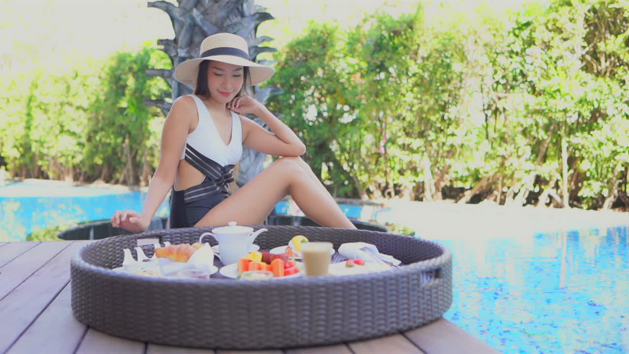 Asian woman tourist pouring morning coffee into the cup. Exotic breakfast tray full of fruits and bread, desserts and drinks near swimming pool in the hotel resort