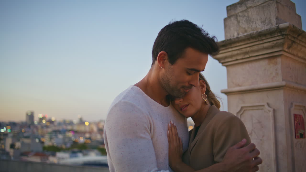 Primo piano di un uomo innamorato che bacia la fronte e abbraccia teneramente una ragazza, con vista sulla città al tramonto.