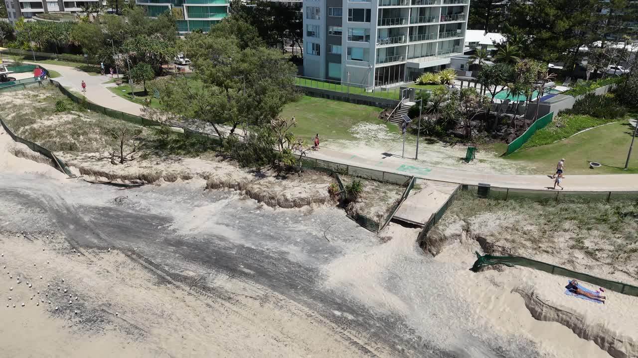 Aerial view of families walking near coastal erosion in Gold Coast, Australia. Bright daylight highlights the sandy beach and surrounding greenery