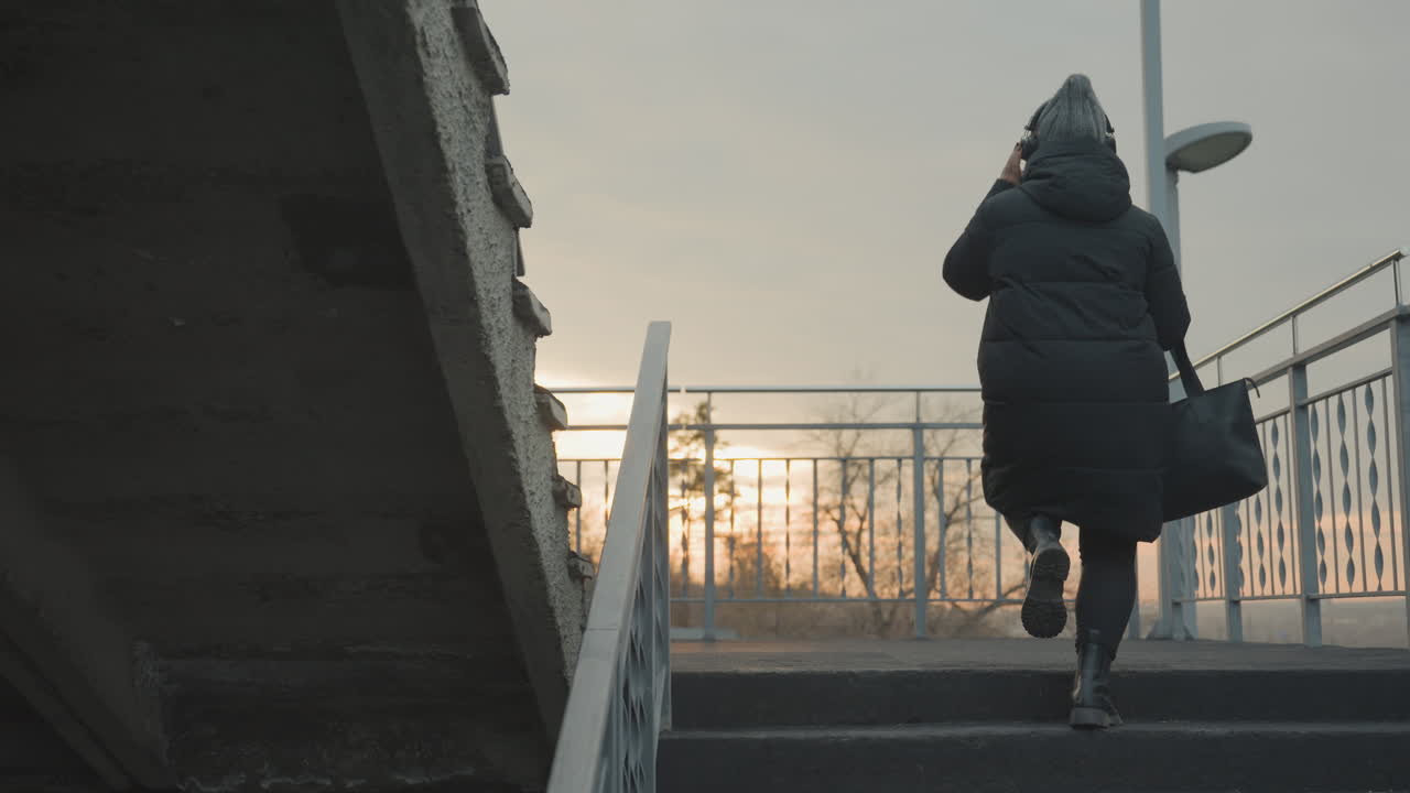 Woman ascending metal stairs at sunrise wearing puffy coat and tote, stepping to internal rhythm and occasional dance sway, city horizon glowing, solitary commuter vibe with calm light