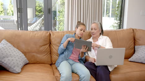 asian grandmother and biracial granddaughter sitting on sofa, using devices