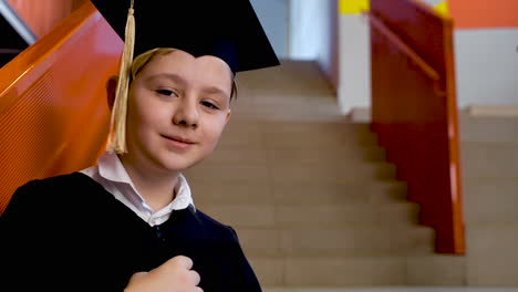 portrait of a happy preschool male student in cap and gown holding graduation diploma and looking at the camera