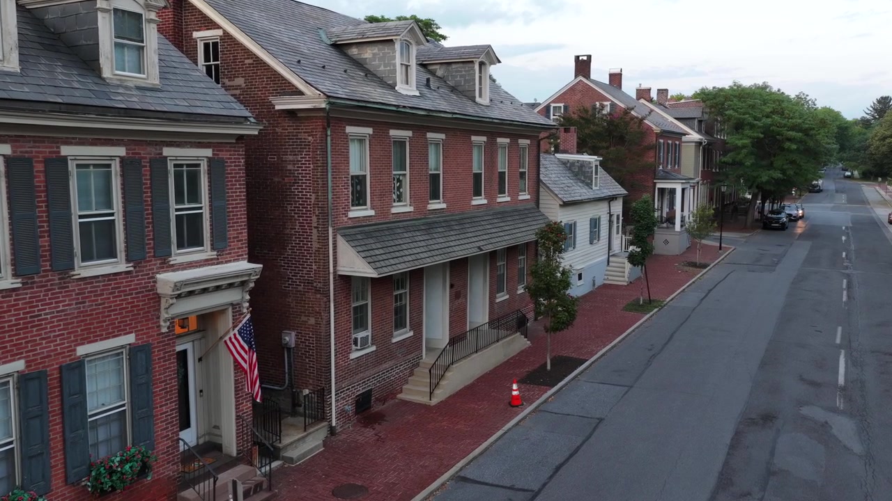 Bandiera americana pende accanto all'ingresso di una casa a schiera in mattoni su una storica strada alberata a Betlemme, Pennsylvania, con abbaini e tetti in ardesia che fiancheggiano il marciapiede lastricato in mattoni sottostante. Vista aerea con drone