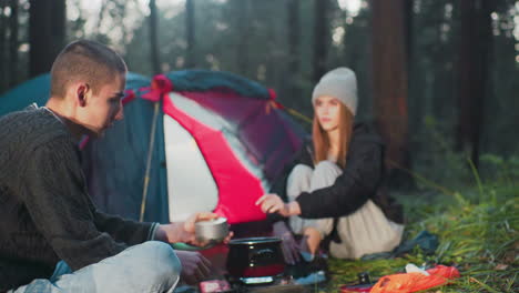 woman in gray beanie and tracksuit sits near tent in forest handing canned food to man as sunlight highlights her face during calm camping moment with boiling pot on portable stove