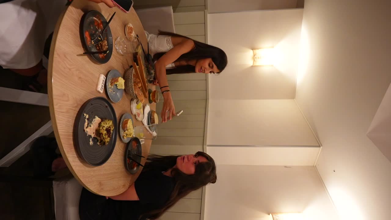 Women shows her phone to a friend while another one uses it during a meal at a restaurant table. Vertical