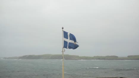 Worn-Scottish-flag-flying-in-strong-stormy-winds-on-remote-Scottish-island-with-fog-behind