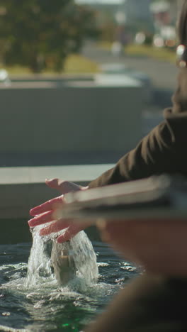 hombre con abrigo negro sentado al aire libre realizando varias tareas con una tableta cerca del agua de manantial bajo la luz del sol brillante, la mano en equilibrio en un gesto activo, con un fondo bokeh suave