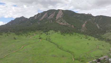 Aerial-view-of-warm-sun-hitting-Boulder-Colorado-Flatiron-mountains-above-Chautauqua-Park-with-full-green-pine-trees,-grass,-and-blue-skies-with-clouds-on-a-beautiful-summer-day-for-hiking