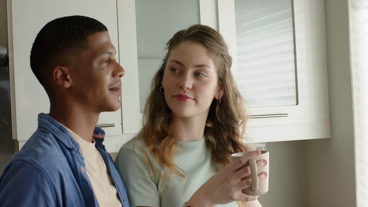 Soft light filtering blinds drawing gaze, woman turning toward man smiling, sipping from coffee mug
