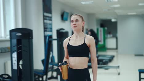 Pretty woman in a black top is drinking a drink from a yellow shaker and walking through the gym on the background of sports equipment. Healthy lifestyle