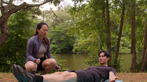 woman teaching man wearing sports clothing in outdoor yoga class surrounded by forest and lake or river 2