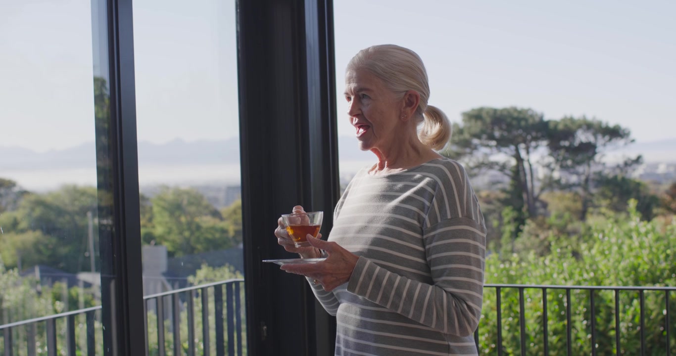 Donna anziana in piedi sul balcone vicino alla porta a vetri, che tiene una tazza con piattino, si ferma, guarda e poi sorseggia.