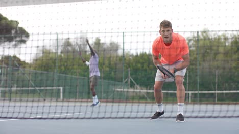 Two-diverse-male-friends-playing-doubles-serving-ball-over-net-at-outdoor-court-in-slow-motion