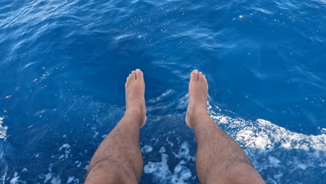 A-pair-of-male-legs-dangling-over-blue-water-from-a-moving-boat