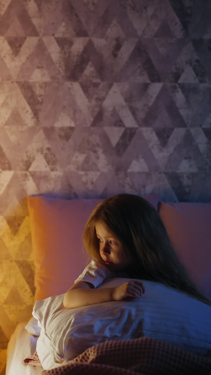 Offended little girl with tears in eyes leaning on large pillow looks at play house with glowing night lamp inside on bed in room in evening slow motion