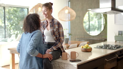 smiling lesbian couple sitting on kitchen counter, enjoying coffee together