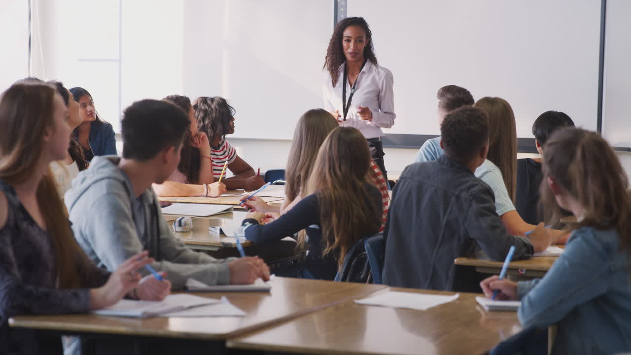 maestra de secundaria haciendo preguntas de pie junto a una pizarra interactiva enseñando una lección